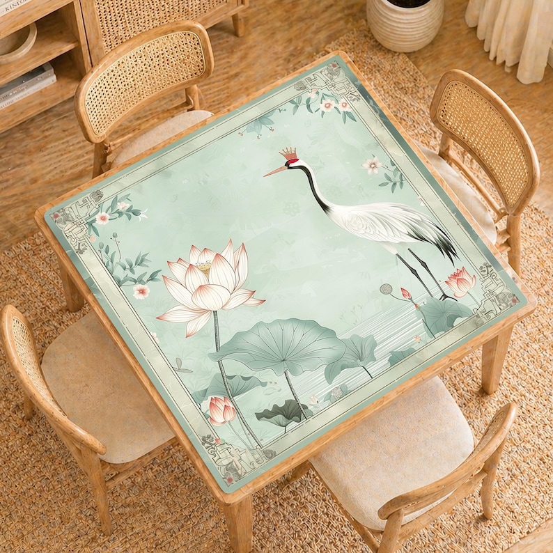 May include: Square wooden table with a light green floral design featuring a crane, lotus flowers, and leaves. The table is surrounded by four wooden chairs with woven backs and beige cushions. The table is set on a woven rug.