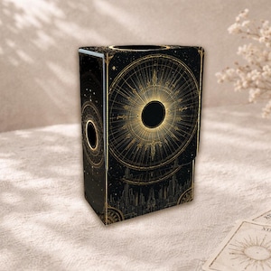 Könnte beinhalten: Schwarze Tarotkartenbox mit goldenem Himmelsdesign. Die Box zeigt eine detaillierte Illustration eines Sonnenaufgangs und einer Stadtlandschaft. Die Box ist rechteckig mit einer kreisförmigen Öffnung oben.