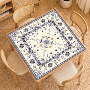 May include: Square wooden table with a floral patterned tablecloth. The tablecloth features a cream background with blue and gold floral designs. Four wooden chairs with woven backs surround the table, creating a dining or living space.