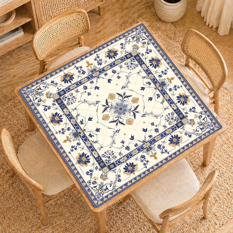 May include: Square wooden table with a floral patterned tablecloth. The tablecloth features a cream background with blue and gold floral designs. Four wooden chairs with woven backs surround the table, creating a dining or living space.