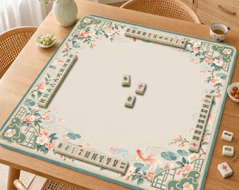 Op de afbeelding: Een vierkante mahjongmat met een crèmekleurig midden en een rand versierd met roze kersenbloesems, groen gebladerte en oranje goudvissen. De mat is gerangschikt met rijen ivoorkleurige mahjongtegels met groene en rode karakters.