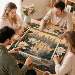May include: A mahjong game set featuring a rectangular play mat with a printed illustration of a city skyline above a lotus pond. The set includes green tile racks and white mahjong tiles with black Chinese characters arranged on the mat.