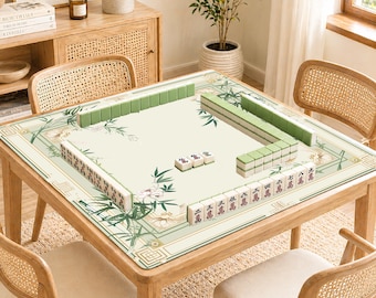Op de afbeelding: Een houten vierkante mahjongtafel met een crèmekleurig speeloppervlak. Het oppervlak heeft een bloemen- en bamboe-ontwerp met groene en witte accenten. Groene mahjongtegels zijn op de tafel gerangschikt, klaar om te spelen. De tafel is omgeven door vier rotan stoelen.