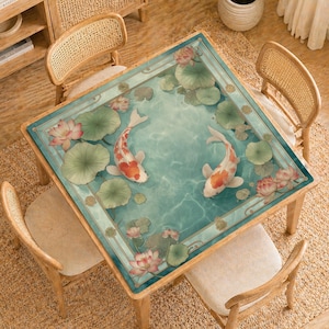 May include: Square wooden table with a decorative top featuring a koi pond design. The design includes two koi fish, lotus flowers, and lily pads in a teal-colored water setting. The table is surrounded by four wooden chairs with woven backs.