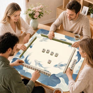 May include: A square mahjong mat featuring a cream-colored surface with a blue koi fish and lotus leaf pattern. The mat includes four green tile racks and a set of white mahjong tiles with black Chinese characters arranged in the center.