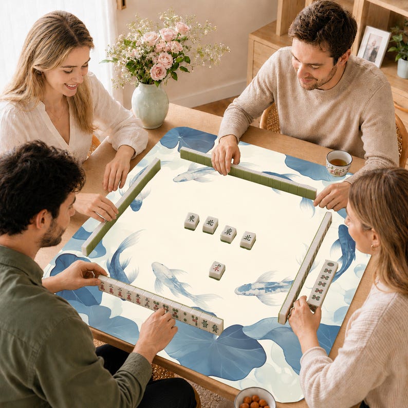 May include: A square mahjong mat featuring a cream-colored surface with a blue koi fish and lotus leaf pattern. The mat includes four green tile racks and a set of white mahjong tiles with black Chinese characters arranged in the center.