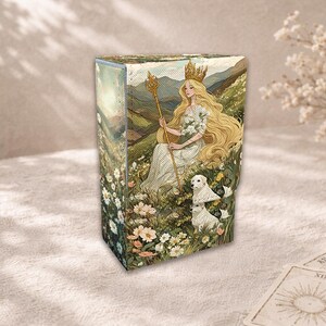May include: A rectangular tarot card box with a serene illustration of a woman with long blonde hair, a crown, and a staff, seated in a field of flowers with two white dogs. The box has a landscape design with a soft, vintage aesthetic.