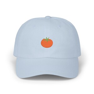 May include: A pale blue baseball cap featuring an embroidered orange tomato with green leaves. The cap has a curved peak and an adjustable fabric strap. The tomato design is centrally placed.