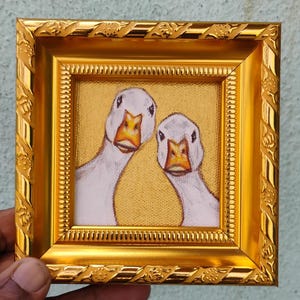 May include: A framed miniature painting featuring two white geese with orange beaks against a gold background. The painting is set within a decorative gold frame with ornate detailing, creating a charming and whimsical artwork.