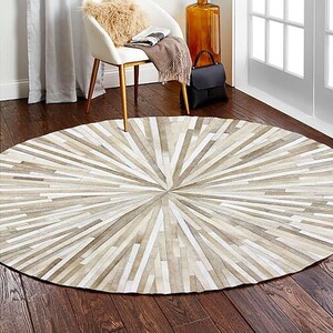 May include: A round rug with a sunburst design. The rug features alternating strips of beige, cream, and light brown, radiating from the center. The rug is made of a natural material and is placed on a dark wood floor.