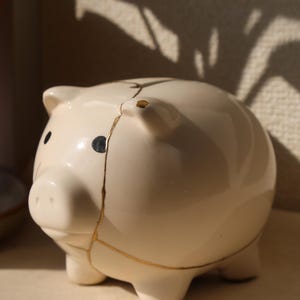 以下が含まれることがあります： 黒い目と鼻を持つ、クリーム色の陶器の貯金箱。 貯金箱は金色の金継ぎで修復され、ひび割れが強調されています。 デザインは、4つの小さな足を持つ、クラシックで丸みを帯びた形状です。