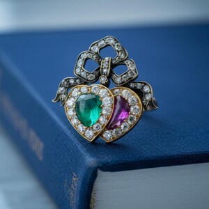 Op de afbeelding: Een vintage ring met twee hartvormige edelstenen, een smaragdgroene en een paarse, omringd door diamanten. De ring, gezet in goud en zilver, ligt op een blauw boek, wat een klassiek sieradenontwerp laat zien.