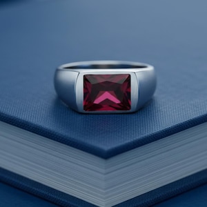 May include: A silver-toned ring featuring a rectangular, deep red gemstone set in a smooth, polished metal band. The ring has a substantial, solid design with a flat top setting that highlights the vibrant, faceted red stone at its center.