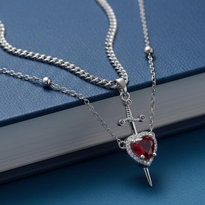 May include: Silver-toned necklace featuring a heart-shaped pendant with a red gemstone center, encircled by smaller clear stones. A sword design is centered above the heart. The necklace is displayed on a blue surface.