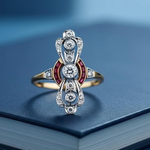 Può includere: Un anello d'oro ornato con un design centrale con un grande diamante rotondo, circondato da diamanti più piccoli e pietre preziose rosse. L'anello è posto su uno sfondo blu, evidenziando i suoi dettagli intricati e la sua maestria.