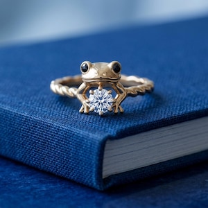 May include: A gold ring with a frog design, holding a clear, round gemstone. The frog has black eyes and sits on a twisted band. The ring is set against a blue backdrop.