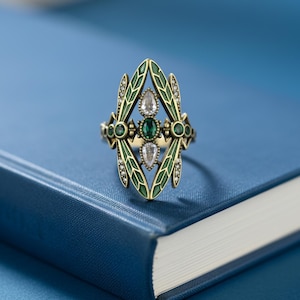 May include: An ornate gold-toned ring with a central emerald-green stone and two clear, teardrop-shaped stones. The ring features green enamel accents and small, clear stones, set against a blue background.