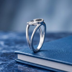 May include: A silver ring with a heart-shaped design. The heart is filled with black stones and surrounded by smaller white stones. The ring is set on a blue surface.