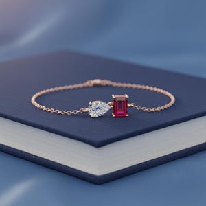 May include: A delicate rose gold bracelet featuring a pear-shaped diamond and a rectangular ruby. The bracelet is set on a dark blue surface, creating a striking contrast. The jewelry piece is a beautiful accessory.