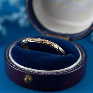 Puede incluir: Un anillo de oro con un diseño floral se muestra dentro de un joyero de terciopelo azul. El anillo tiene un delicado patrón de pequeñas flores y líneas. La caja es redonda con un exterior azul oscuro y un cierre dorado.