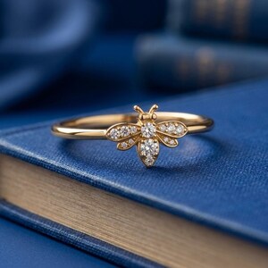 May include: A gold-colored ring with a bee design. The bee's body and wings are adorned with small, sparkling gemstones. The ring is set against a blue background, resting on a book.