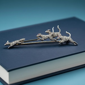 Op de afbeelding: Zilveren broche met een ontwerp van rennende honden. De broche toont vier honden, elk versierd met kleine, sprankelende stenen en rode edelsteenogen. De honden zijn gerangschikt op een goudkleurige staaf.