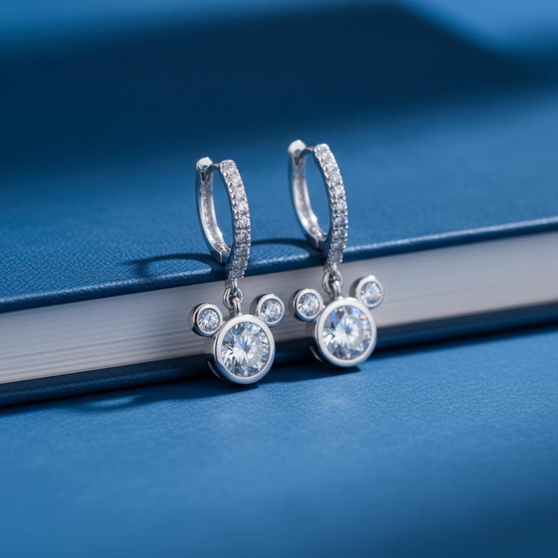 K&ouml;nnte beinhalten: Ein Paar silberfarbene Creolen-Ohrringe mit einem Design, das von Mickey Mouse inspiriert ist. Jeder Ohrring hat einen gro&szlig;en, runden, klaren Edelstein in der Mitte, umgeben von kleineren Edelsteinen und einem kleineren Edelstein auf jeder Seite.