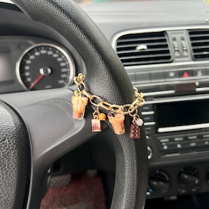 May include: A gold-colored charm bracelet with miniature drink and food charms, including a boba tea, hangs from a black steering wheel. The car's dashboard and speedometer are visible in the background, with a focus on the bracelet's decorative elements.