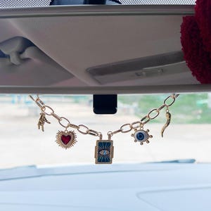 May include: A gold-toned metal chain hanging from a rearview mirror, featuring five charms: a hand gesture, a red heart with rays, a rectangular blue plaque with an eye design, a round blue eye charm, and a curved horn-shaped pendant.