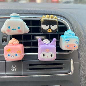 May include: Five square-shaped car air vent clips featuring cartoon character designs. The clips include a white dog, a black penguin with a gold crown, a pink cube with a star-topped head, a purple character with a skull, and a blue cube with stars.