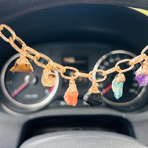 Op de afbeelding: Een goudkleurige ketting met meerdere bedels van ruwe, ongeslepen edelstenen in diverse kleuren zoals bruin, oranje, zwart, blauw, paars en roze, elk voorzien van een goudkleurige metalen zetting die aan de ovale schakels van de ketting hangt.