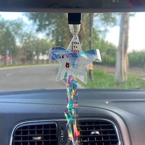 Può includere: Un ornamento a forma di stella per lo specchietto retrovisore dell'auto con una finitura traslucida e iridescente e piccole perline dorate incastonate. Include un nastro bianco per appenderlo e lunghi nastri olografici scintillanti.