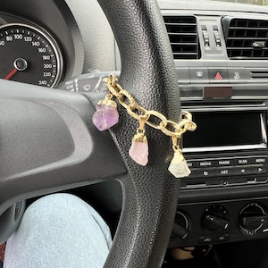 以下が含まれることがあります： 車のハンドルにぶら下がっている、3つの宝石チャームが付いたゴールドカラーのチェーン。宝石は紫、ピンク、白など様々な色があります。車のダッシュボードとハンドルが見えます。
