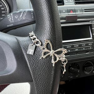 Puede incluir: Un dije de mariposa plateada con una cadena y una pequeña etiqueta rectangular con la letra "T" cuelga de un volante negro. La mariposa tiene intrincados detalles recortados. El salpicadero del coche es visible en el fondo.