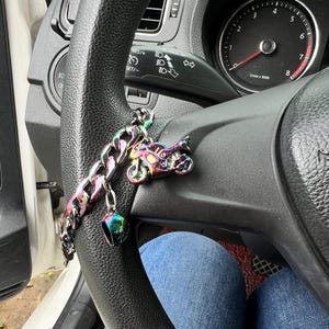 May include: A motorcycle-themed keychain hanging from a black steering wheel. The keychain features a small, iridescent motorcycle charm, a matching helmet charm, and a chunky chain. The steering wheel is in a car interior.