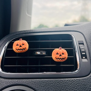 Könnte beinhalten: Zwei kleine, orangefarbene, texturierte Jack-o'-Lantern-Kürbis-Lüftungsclips, die an den horizontalen schwarzen Lamellen einer Fahrzeuglüftung befestigt sind. Jeder Kürbis hat ein geschnitztes Gesicht mit dreieckigen Augen und einem gezackten Mund.