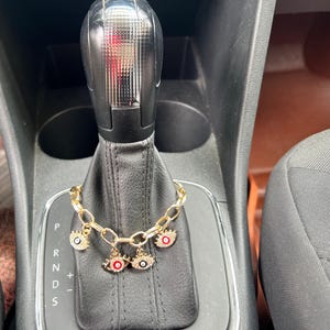 Op de afbeelding: Een goudkleurige schakelarmband met drie oogvormige bedels. Elke bedel is ingelegd met kleine doorzichtige steentjes en heeft een centrale pupil van rode of zwarte emaille. De armband hangt over een zwarte lederen versnellingspook van een auto.