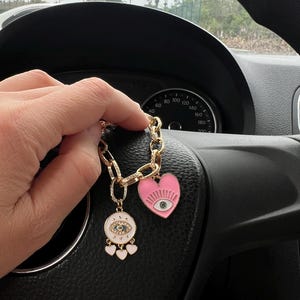 Op de afbeelding: Goudkleurige sleutelhanger met een schakelketting ontwerp. De sleutelhanger heeft een roze hartbedel met een oogontwerp en een witte cirkelvormige bedel met een oogontwerp en drie kleine roze hartbedels. De sleutelhanger wordt voor het stuur van een auto gehouden.