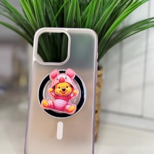 May include: A clear phone case with a pink border and a pop-up grip featuring a cartoon bear in a pink outfit. The phone case is set against a background of green plants.