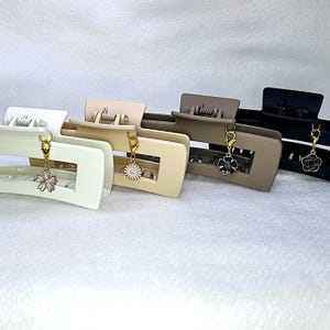 May include: Four rectangular hair claw clips in white, beige, taupe, and black. Each clip has a gold-colored charm with a flower design. The clips are arranged in a row, showcasing their different colors and styles.