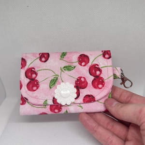 May include: A small, pink fabric wallet with a cherry pattern. The wallet has a white flower embellishment and a silver-colored clasp. The cherry design features red cherries and green leaves on a light pink background.