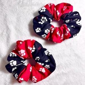 May include: Two scrunchies with a red and navy blue paw print design. The scrunchies are made of fabric and are designed to hold hair. The paw prints are white.
