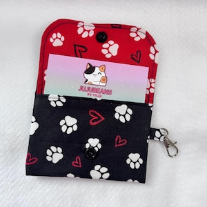 May include: Open fabric card holder with a black and white paw print pattern on the exterior and a red paw print pattern on the interior. The card holder has a snap closure and a silver-colored clasp. A business card with a cat logo is visible.