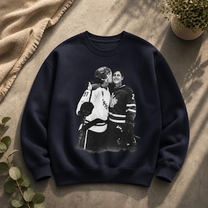 May include: Navy blue sweatshirt featuring a black and white image of two hockey players kissing. One player wears a white jersey with the number 91, the other a dark jersey with the number 2. The sweatshirt is a crew neck.