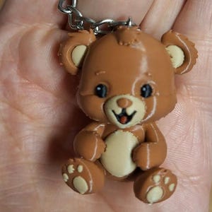 Op de afbeelding: Een bruine teddybeer sleutelhanger met een zilveren metalen ring. De beer heeft een lichtbruine buik en binnenoren, met zwarte ogen en een rode tong. De voeten van de beer hebben details van de voetzool.