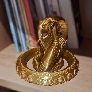 Könnte beinhalten: Eine goldene Kobra-Statue mit detailliertem, strukturiertem Design. Der Kopf der Schlange ist erhoben, die Zunge ausgestreckt. Die Statue steht auf einer Holzoberfläche, mit einem verschwommenen Hintergrund aus Büchern.