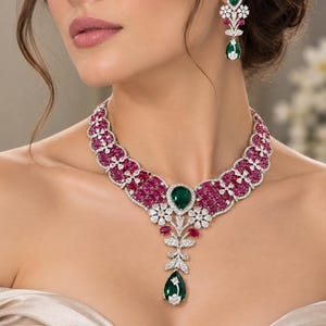 Op de afbeelding: Een sieradenset bestaande uit een ketting en bijpassende oorbellen. De ketting is voorzien van ovaal geslepen rode stenen, heldere witte stenen en een centrale peervormige groene steen in een zilverkleurige zetting.