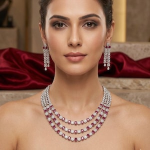 Puede incluir: Una mujer lleva un collar de tres hilos y pendientes a juego. La joyería presenta una combinación de piedras blancas y piedras preciosas rojas. El collar y los pendientes se colocan sobre un fondo rojo.