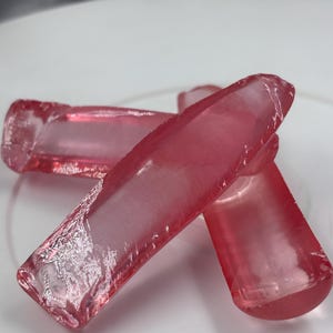 May include: Three translucent, elongated, cylindrical objects with a vibrant pink hue. Each piece features a smooth, polished surface with subtle internal variations and rounded ends, resembling glass or resin rods arranged in a cluster.