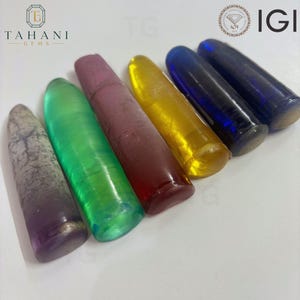 Puede incluir: Seis especímenes de piedras preciosas cilíndricos y alargados dispuestos en fila. Los colores incluyen un gris-púrpura moteado, un verde translúcido vibrante, un rojo granate profundo, un amarillo brillante, un azul real profundo y un azul marino oscuro.